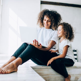 Cheerful ethnic curly woman showing to cheerful daughter laptop screen while sitting in white room in front of window at daytime